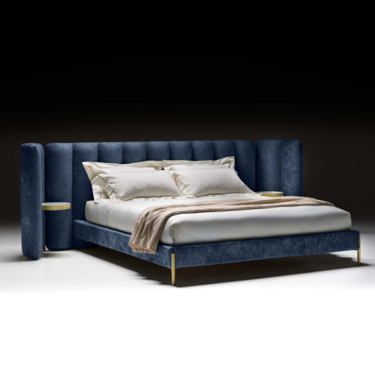 The Contemporary Bed With Winged Wide Headboard offers a luxurious touch with its blue upholstered headboard and side panels, accented by gold details and built-in nightstands. It's neatly arranged with white linens and pillows, complemented by a beige throw blanket at the foot of the bed, all set against a black background.