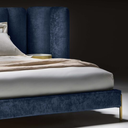 Contemporary Bed With Winged Wide Headboard A close-up view of the Contemporary Bed With Winged Wide Headboard, showcasing its dark blue headboard and frame. The bed is dressed in white bedding with a beige blanket casually draped across it. In the background, the corner of a round, gold-colored bedside table can be seen.
