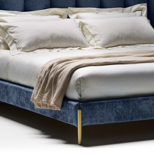 Contemporary Bed With Winged Wide Headboard A neatly made bed with white sheets, four white pillows, and a light beige throw blanket draped across the foot. The Contemporary Bed With Winged Wide Headboard is upholstered in dark blue fabric and features slender brass-colored legs. Soft lighting highlights the luxurious bedding.