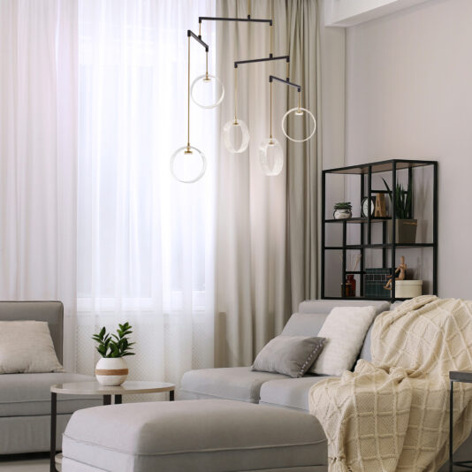 Contemporary Glass Ring Chandelier A modern living room with light grey sofas, a cozy beige throw blanket, and a small round coffee table holding a potted plant. A geometric black shelving unit with decor items stands against the wall, while Contemporary Glass Ring Chandeliers accentuate the space.