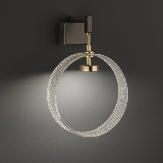 Mounted on a dark background, the Contemporary Glass Wall Light showcases a sleek, circular design. It features a brass-colored fixture paired with a textured glass ring that emits soft, diffused light, casting an elegant glow around the surrounding area.
