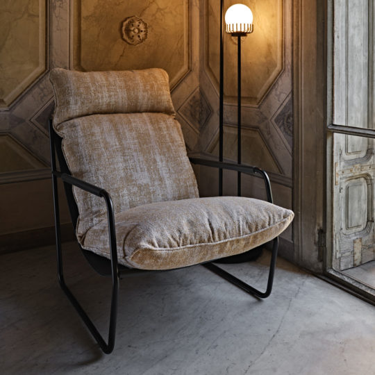 A Contemporary Mid Century Style Metal Armchair with a brownish fabric cushion seat and backrest is placed beside a tall floor lamp with a round, glowing light. This stylish piece sits in a room featuring an ornate, classical wall design and a large window with intricate wooden frame details.