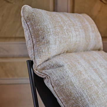 A close-up view of the Contemporary Mid Century Style Metal Armchair featuring a matte black metal frame and plush beige cushions with a subtle textured pattern. The background shows a blurred wooden panel wall, emphasizing the armchair's contemporary mid-century style and comfort.