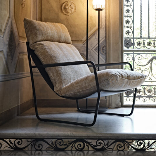 Contemporary Mid Century Style Metal Armchair A stylish **Contemporary Mid Century Style Metal Armchair** with cushions and a sleek black frame is placed on a marble platform in a room featuring ornate wall designs and an intricate wrought-iron window railing. Beside the armchair, there stands a tall modern floor lamp with a ribbed shade.