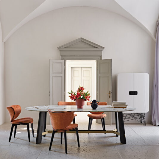 Contemporary Oval Quartz Dining Set A contemporary dining room showcases the elegant Contemporary Oval Quartz Dining Set, complete with four orange upholstered chairs. The table is adorned with a bouquet of red flowers in a vase, a small black sculpture, and several stacked books. The room features arched ceilings, a doorway, and a minimalist white cabinet.