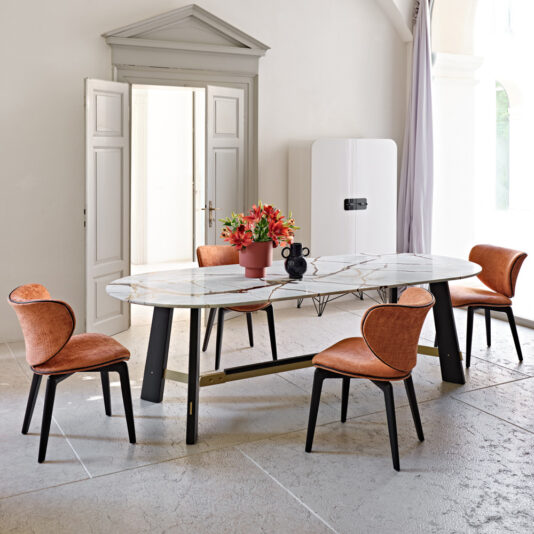 A modern dining room with a Contemporary Oval Quartz Dining Set featuring a marble-top table and four orange chairs with black legs. The table has a centerpiece with vibrant flowers. The room showcases tall white double doors, a minimalist white fridge, and a tall arch window with a curtain.