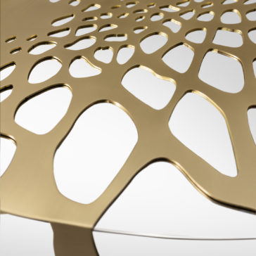 A close-up of the Exclusive Cut Out Round Coffee Table reveals its gold-colored, intricately designed metal surface with abstract, organic cut-out patterns reminiscent of interconnected cells or a honeycomb structure. The table's exclusive design stands out beautifully against a plain, neutral white background.