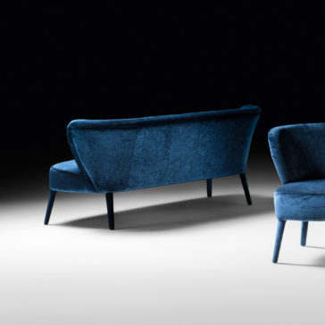 High End Mid Century Style Day Sofa A stylish blue velvet loveseat and a matching High End Mid Century Style Day Sofa, embodying a high-end mid-century style, are set against a black and white gradient background. The loveseat is positioned at an angle, showcasing its sleek design and slim legs, while the day sofa chair is partially visible in the foreground.