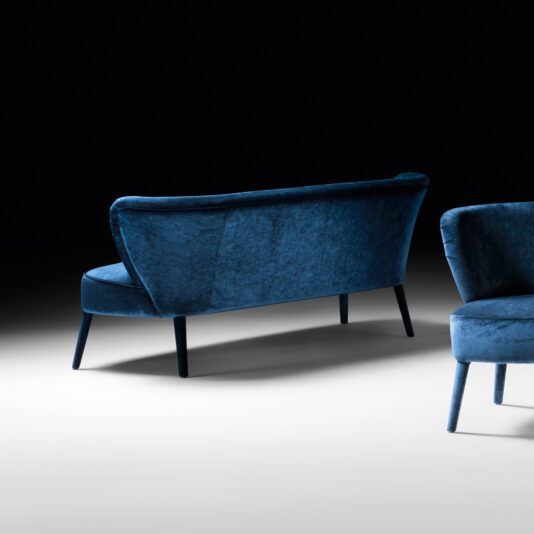 High End Mid Century Style Day Sofa A stylish blue velvet loveseat and a matching High End Mid Century Style Day Sofa, embodying a high-end mid-century style, are set against a black and white gradient background. The loveseat is positioned at an angle, showcasing its sleek design and slim legs, while the day sofa chair is partially visible in the foreground.