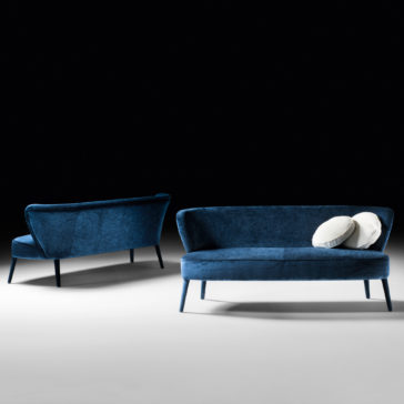 High End Mid Century Style Day Sofa Against a black background, two blue velvet upholstered sofas are showcased. The High End Mid Century Style Day Sofa is positioned to display its front and right side, adorned with two grey cushions. The other sofa is oriented to highlight its back and left side.