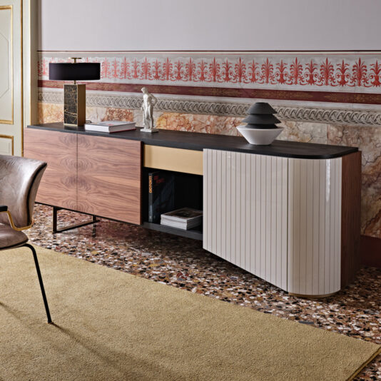 A High End Modern Canaletto Walnut Sideboard, showcasing a sophisticated blend of wooden and glossy white finishes, stands against a wall adorned with ornate floral wallpaper. The sideboard is adorned with decorative items such as books, a lamp, and a ceramic vase. Beside it sits a leather chair on a patterned floor complemented by a beige rug.