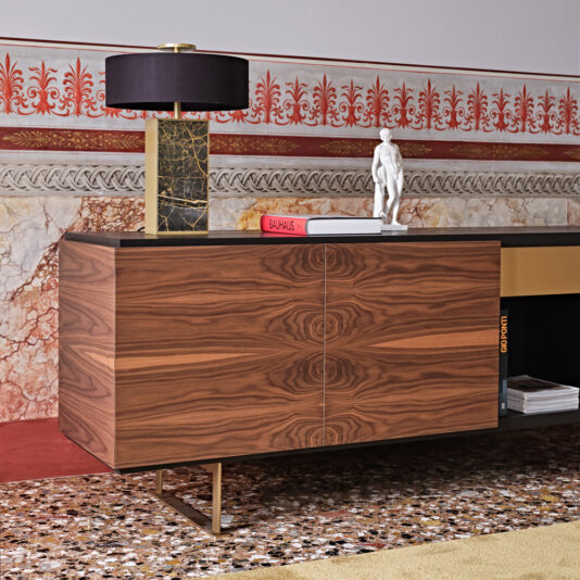 A High End Modern Canaletto Walnut Sideboard with a pristine wood finish stands against a decorated wall adorned with red intricate patterns and marble sections. Atop the sideboard, there is a black and gold lamp, a small white statue, and a red book. The floor showcases a speckled tile design.