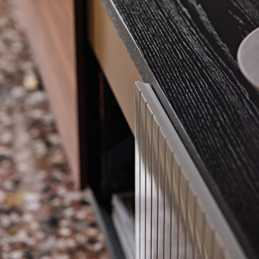 A close-up of the High End Modern Canaletto Walnut Sideboard against a textured wall. The sideboard boasts a luxurious Canaletto walnut surface on top, a metallic front with elegant vertical lines, and a partially visible open shelf. The floor beneath features a speckled pattern.