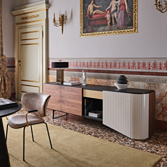 A stylish interior room featuring a gold-trimmed door, ornate wall sconces, and a High End Modern Canaletto Walnut Sideboard with a white curved cabinet door. A modern black lamp and decorative vase sit atop the sideboard, while a framed classical painting hangs above it. A beige carpet covers the patterned floor.