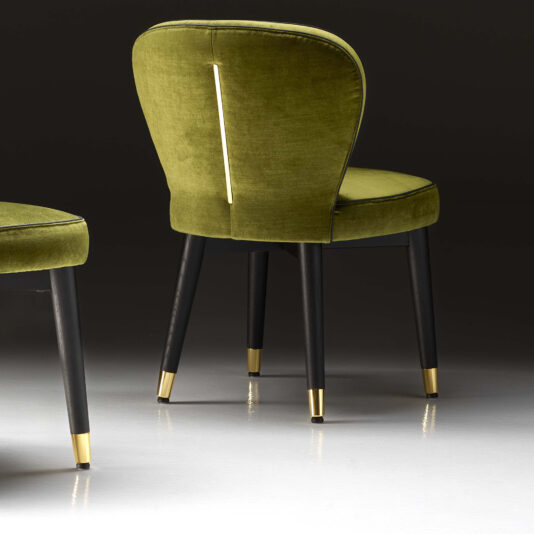 The High End Velvet Dining Chair, upholstered in elegant green velvet, showcases a curved backrest with a striking gold-colored metal accent at its center. It stands on dark wooden legs adorned with gold tips. To the left, another similar chair is partially visible, while the dark background accentuates its luxurious details.