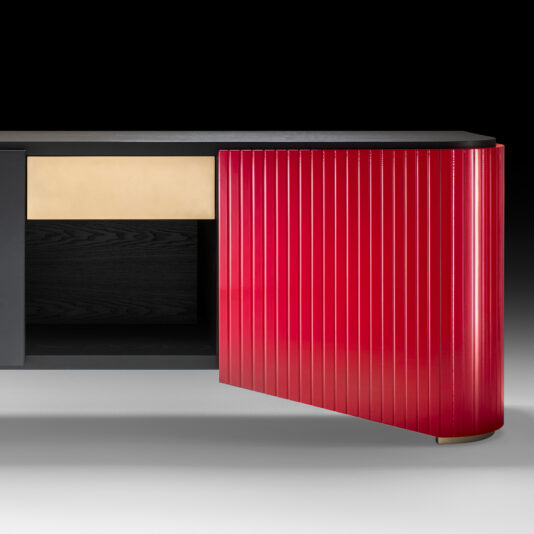 The Large Contemporary Sideboard showcases a sleek and modern design, featuring a black top surface, a luxurious golden drawer, and an eye-catching red door with vertical ribbing on one side. The door is partially open, revealing an empty interior space inside this statement piece of furniture. It stands boldly on a light grey floor with a black backdrop.