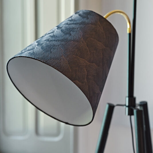 Large Modern Studio Style Floor Lamp A close-up shot of the Large Modern Studio Style Floor Lamp featuring a textured dark grey lampshade, angled slightly downward. The lamp's gold neck and tripod base are prominently displayed. In the background, a softly lit window is out of focus, adding a touch of natural light to the scene.