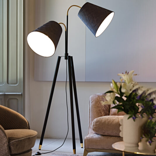 A stylish Large Modern Studio Style Floor Lamp with two illuminated black lampshades, each directed in opposite directions, stands on a three-legged tripod base in a cozy living room. The setting includes a beige armchair, a wooden panel wall, and a table with a vase of flowers.