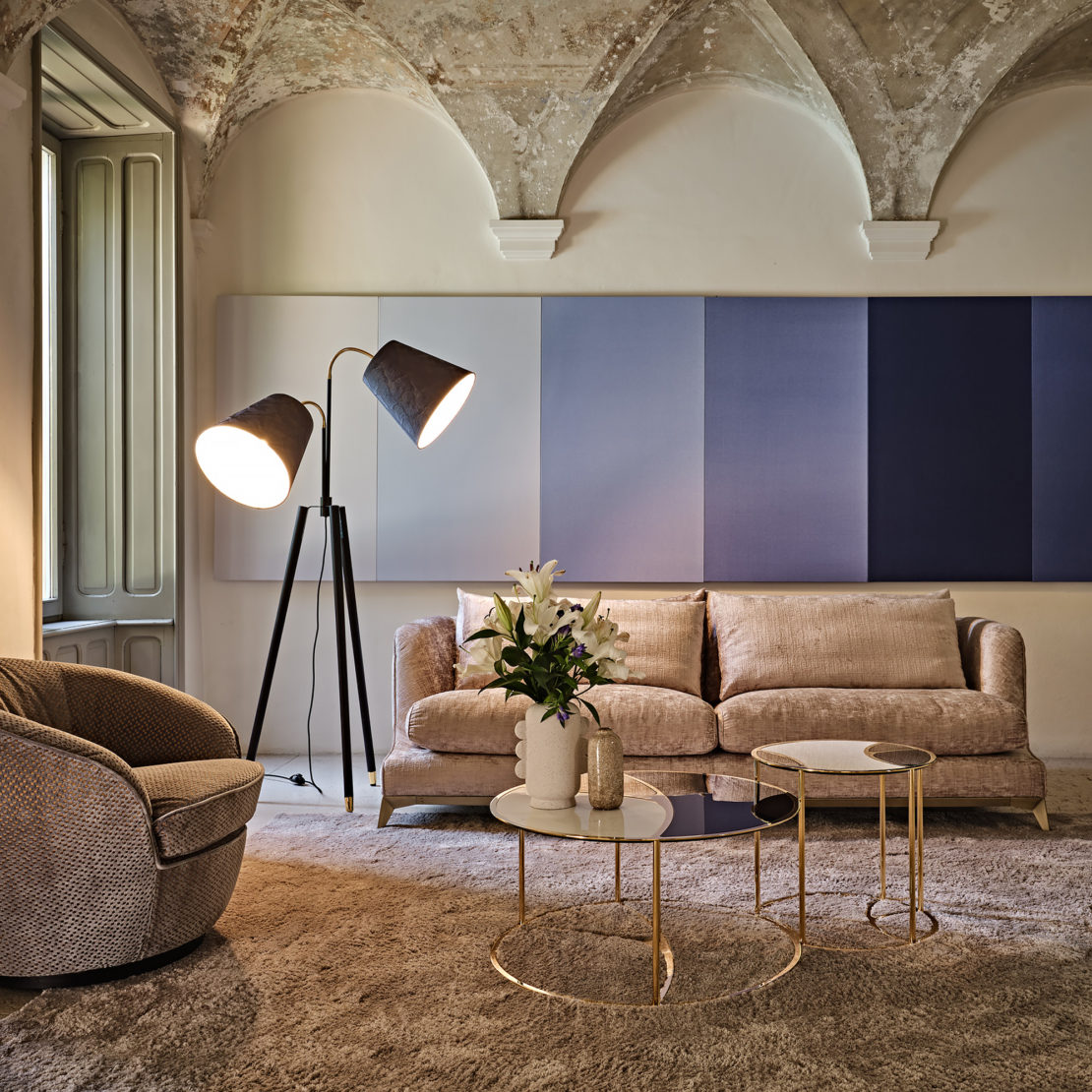 A cosy living room featuring a beige sofa, a round armchair, and the Large Modern Studio Style Floor Lamp. The space boasts arched ceilings, a multi-colored wall panel, light wooden furniture, and decorative items on circular tables. Natural light filters through a large window on the left.