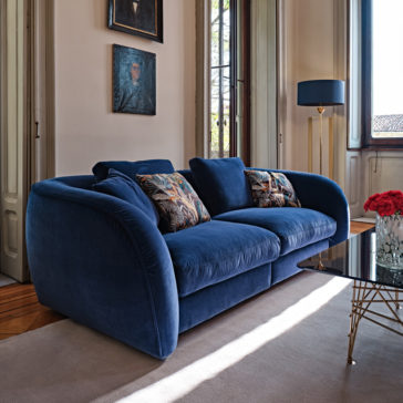 Large Modern Velvet Modular Sofa A sleek modern living room features a Large Modern Velvet Modular Sofa in deep blue, adorned with two printed throw pillows. Above the couch, two framed portraits are mounted on a neutral-colored wall. A glass coffee table with gold legs holds a vase of red flowers, while a tall floor lamp stands nearby.