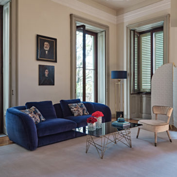 Large Modern Velvet Modular Sofa A stylish living room with large windows, a Large Modern Velvet Modular Sofa in deep blue, a modern glass coffee table with a gold metal base, and a beige armchair. The table has a vase of red flowers. There are two framed portraits on the wall and a standing lamp.
