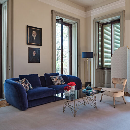 A stylish living room with large windows, a Large Modern Velvet Modular Sofa in deep blue, a modern glass coffee table with a gold metal base, and a beige armchair. The table has a vase of red flowers. There are two framed portraits on the wall and a standing lamp.