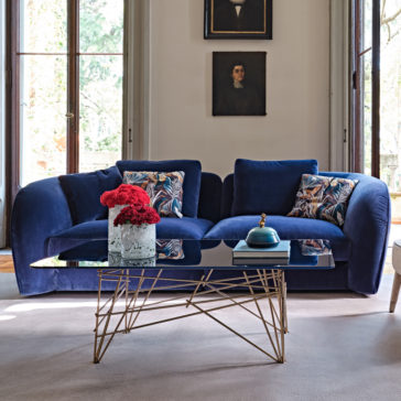 Large Modern Velvet Modular Sofa A modern living room featuring a Large Modern Velvet Modular Sofa adorned with patterned pillows, situated against tall windows. A glass coffee table with a gold geometric base holds a marble vase with red flowers and a stack of books. Two framed portraits hang on the wall behind the sofa.