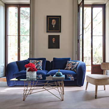 Large Modern Velvet Modular Sofa A modern living room features a luxurious Large Modern Velvet Modular Sofa adorned with colorful cushions, a glass coffee table with a gold geometric base topped with a vase of red flowers and a small decorative piece, large windows, and two framed portraits on the wall.