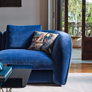 Large Modern Velvet Modular Sofa The Large Modern Velvet Modular Sofa in a rich blue hue is positioned beside a glass coffee table with books on it. The sofa features a vibrant patterned cushion. Adjacent is a large wooden-framed glass door that opens to a lush green outdoor area, letting in natural light.