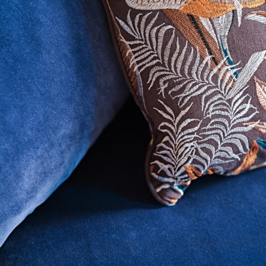 A close-up of a decorative pillow with an intricate leafy pattern in shades of brown and orange, placed on the Large Modern Velvet Modular Sofa in rich royal blue.