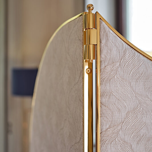 Luxury 3 Leaf Dressing Screen A close-up of the Luxury 3 Leaf Dressing Screen showcases an intricate partition adorned with luxurious, wave-patterned beige fabric and polished gold metal trim. The detailed design emphasizes the screen's elegant craftsmanship and sophisticated aesthetic. In the background, a lamp and window are softly blurred.