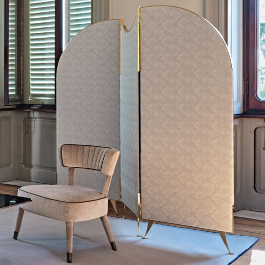 A Luxury 3 Leaf Dressing Screen, featuring light-colored textured fabric, stands next to a beige velvet chair with wooden legs. The scene is set in a room adorned with ornate wooden panels and large windows framed by green shutters, with a neutral rug covering the floor.