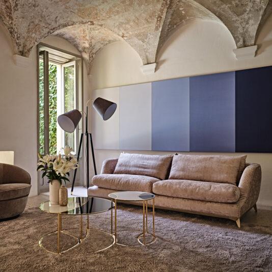 Luxury Contemporary Sofa A modern living room with arched ceilings features a Luxury Contemporary Sofa, a tall floor lamp with dual shades, and two nested glass coffee tables framed in gold. One of the tables displays a vase filled with white flowers. The room boasts large windows that showcase lush greenery outside.