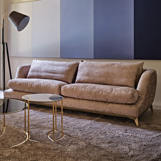 Luxury Contemporary Sofa A modern living room features the Luxury Contemporary Sofa with plush beige cushions, positioned against a wall adorned with sound-absorbing panels in neutral and blue tones. In front of the sofa are two nested round coffee tables with gold frames and black glass tops. A black floor lamp stands nearby.
