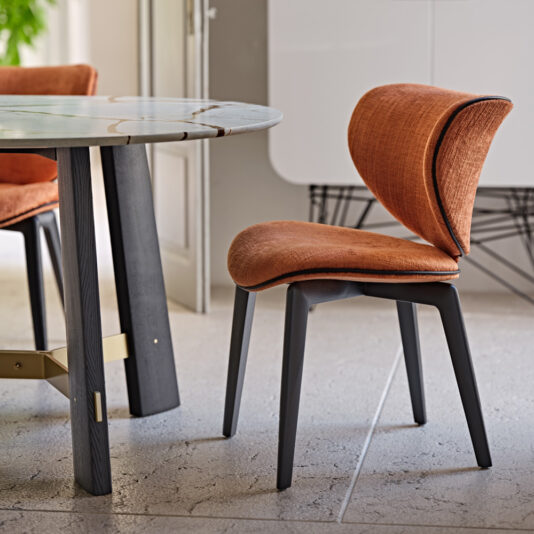 A Mid Century Style Dining Chair with a curved orange upholstered seat and backrest is positioned next to a round marble table featuring black legs and a brass crossbar on a stone tile floor. The arrangement is set in a contemporary dining area that exudes a light and airy ambiance.