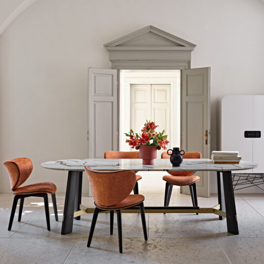 A contemporary dining room featuring a marble-top table with black legs. The table is surrounded by four cushioned Mid Century Style Dining Chairs in orange, each with elegantly curved backs. At the center of the table sits a vibrant red flower arrangement. Double doors and a sleek white cabinet serve as the background.