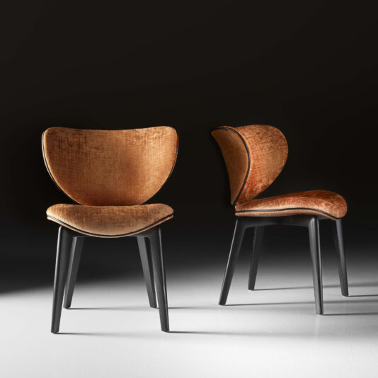 A pair of Mid Century Style Dining Chairs, featuring modern and stylish designs, with dark wooden legs and plush, golden-brown upholstered seats and backs. One chair is displayed from the front view while the other showcases a side profile, all set against a shadowed, minimalist background.