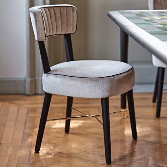 The Modern Designer Velvet Dining Chair features a contemporary design with a round beige velvet cushion seat and backrest adorned with vertical stitching, supported by four sleek black legs connected by an elegant gold crossbar. It is placed on a polished wooden floor beside a dining table with a checkered tabletop.