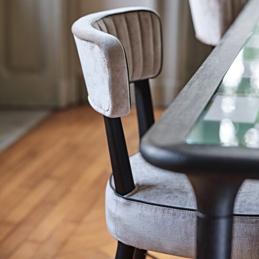 Modern Designer Velvet Dining Chair The Modern Designer Velvet Dining Chair, featuring soft gray velvet upholstery and black legs, is artfully positioned beside a glass-top table. The chair boasts a curved backrest with vertical stitching, all set against the backdrop of wooden flooring and a partially visible door.
