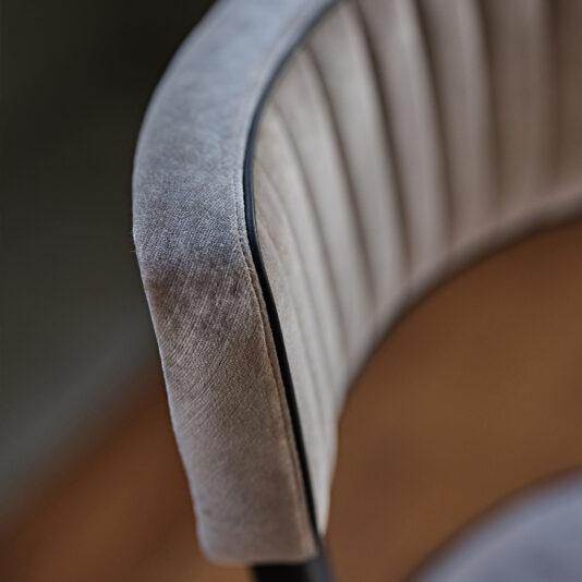 Modern Designer Velvet Dining Chair Close-up of a Modern Designer Velvet Dining Chair with a curved backrest. The chair's fabric is a textured, soft grey velvet with vertical stitching details. The edge is trimmed with a thin black line, and the background is softly blurred.