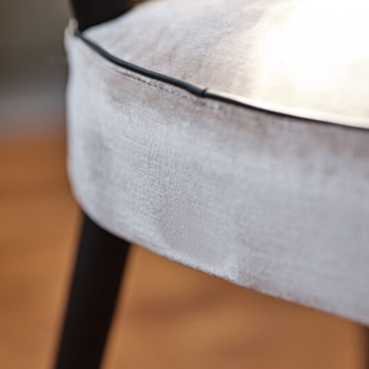 Modern Designer Velvet Dining Chair Close-up of the Modern Designer Velvet Dining Chair, showcasing its light gray velvet upholstery with black piping detail along the edge. The chair's black legs are partially visible, set against a softly blurred background that accentuates the sleek dining chair's design.