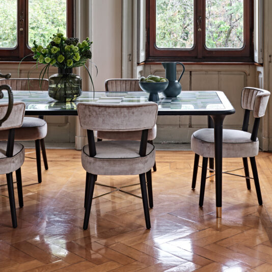 Modern Designer Velvet Dining Chair A dining room with a glass-topped table, surrounded by five Modern Designer Velvet Dining Chairs with wooden legs. The table features a dark vase with green flowers, bowls with green apples, and decorative blue vases. Large windows offer a view of lush greenery outside.