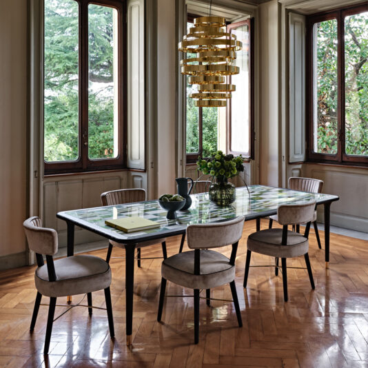 Modern Designer Velvet Dining Chair A dining room with a modern rectangular table set for six, featuring a glass top adorned with a green checkered pattern. Surrounding the table are six Modern Designer Velvet Dining Chairs in gray, each cushioned for comfort. Above, a gold-tiered chandelier adds elegance while large windows reveal views of trees outside, allowing natural light to enhance the room's modern designer aesthetic.