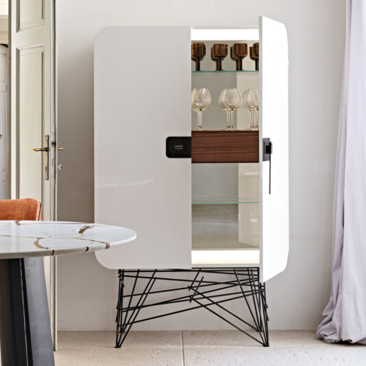 Modern Lacquered Cabinet The Modern Lacquered Cabinet, featuring black metal legs and a partially open door that reveals several glasses on wooden shelves inside, stands next to a round marble-top table. The sleek cabinet design is placed against a light-colored wall with a curtain to the right.