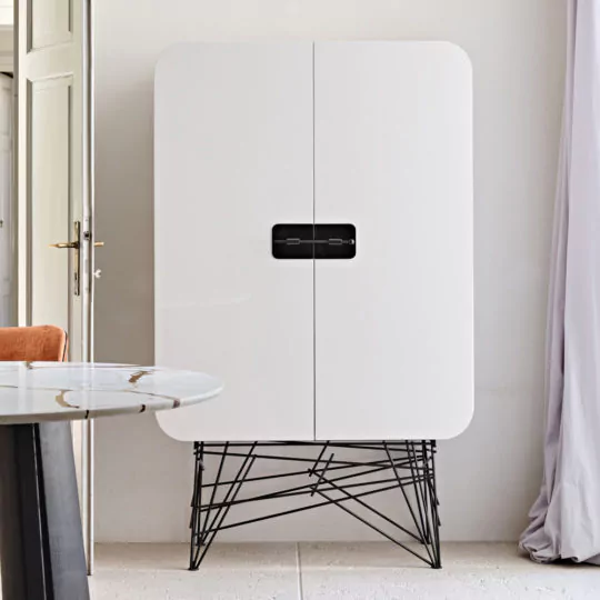 The Modern Lacquered Cabinet, showcasing a minimalist white finish with rounded edges and black metal legs, is situated in a corner adjacent to a curtain. The cabinet boasts a distinctive rectangle handle and rests on an elegant wireframe base. An orange chair and a segment of a round marble-topped table are partially visible nearby.