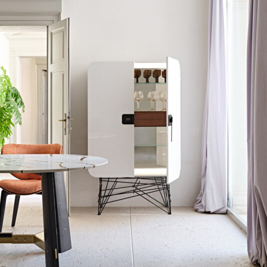 Modern Lacquered Cabinet A Modern Lacquered Cabinet, with one door partially open to showcase wine glasses inside, stands in a bright room adorned with a glass table, an orange chair, a large green plant to the left, and a sheer curtain to the right. The tiled floor reflects the sunlight that fills the space.