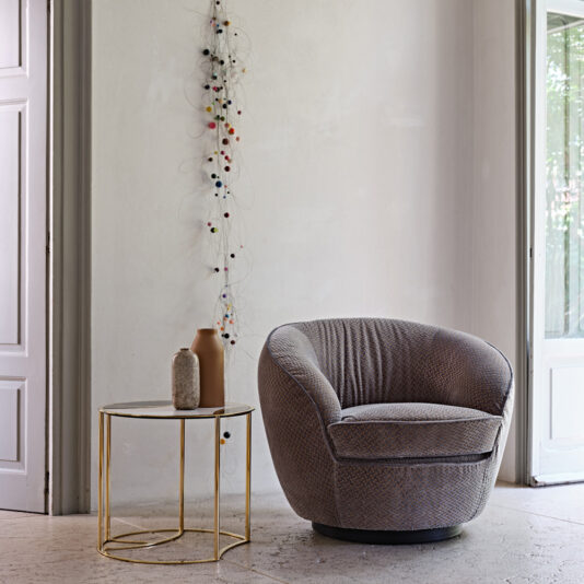 A cozy, rounded Modern Luxury Swivel Armchair in gray is positioned next to a small, elegant side table with a gold frame and glass top. On the table are two decorative vases. The background features a minimalist wall with a string of multicolored ornaments hanging vertically, exuding modern luxury.