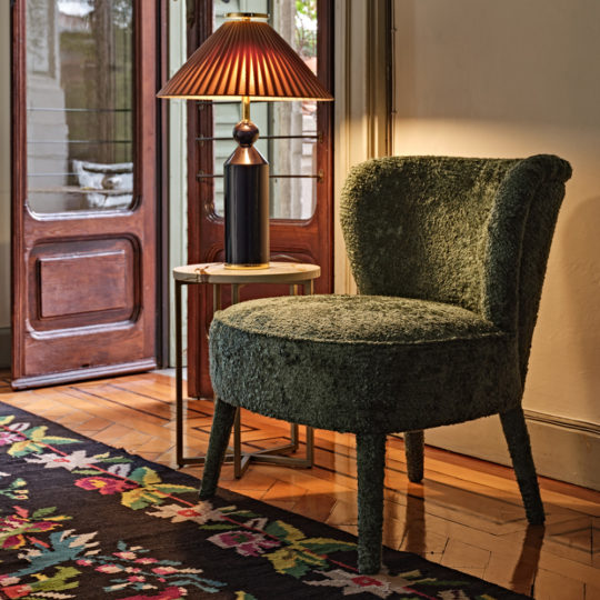 A cozy living room corner showcases the Modern Occasional Chair with a green textured finish, placed beside a small round marble table. A black lamp with a red and gold shade sits on the table, casting a warm glow. The room features wooden floors, a colorful rug, and a door with glass panes.