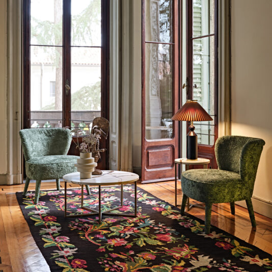 A cozy, elegant living room featuring two Modern Occasional Chairs in green velvet, a white marble coffee table adorned with a flower vase, a vibrant floral-patterned rug, and a small stand topped with a red lampshade. Large windows and a glass door open to reveal an inviting outdoor view.