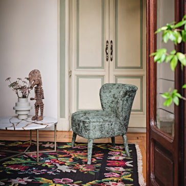 Modern Occasional Chair A cozy corner featuring a textured, green Modern Occasional Chair on a colorful, patterned rug. A marble-top side table holds a potted plant and a wooden sculpture. Pale, ornate double doors in the background and greenery peeking in from the right complete the scene.